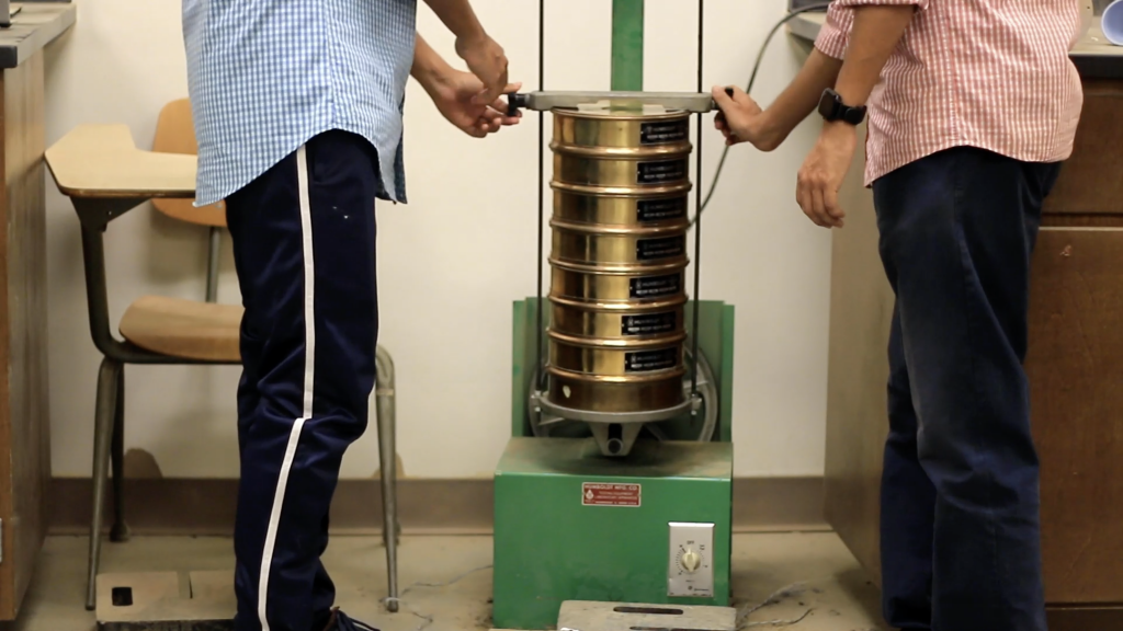 Two persons are putting a stack of sieve into the sieve shaker