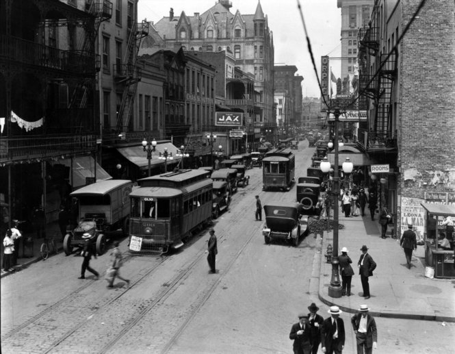 A black and white photo from 1920 shows men in suits and hats, early automobiles, and trolley cars on tracks down the middle of the St. Charles Street, New Orleans.