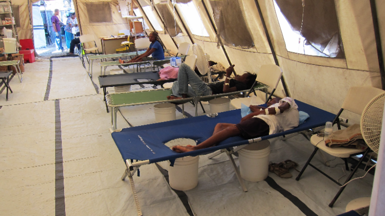 A sparse room contains five cots lining the right side of the room and an opening to the outside at the far side of the room. Four people are lying on the cots. The person toward the front of the photo is wearing bandages on one arm, a white shirt and black shorts. A person in the next cot is wearing a black shirt and gray pants.