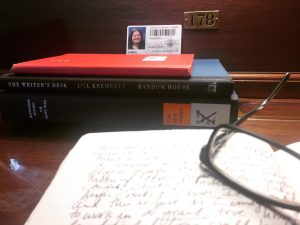 Close up of a desk in the Library of Congress. An open journal with writing on the pages is in the foreground with glasses on top. In the background is a stack of books with a library card resting on top.