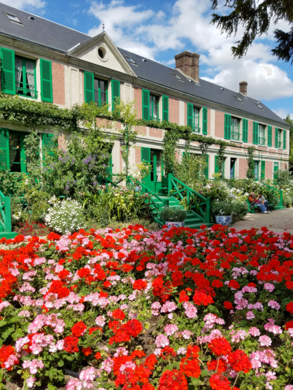 Red and pink geraniums fill a flowerbed in front of a 2-story house